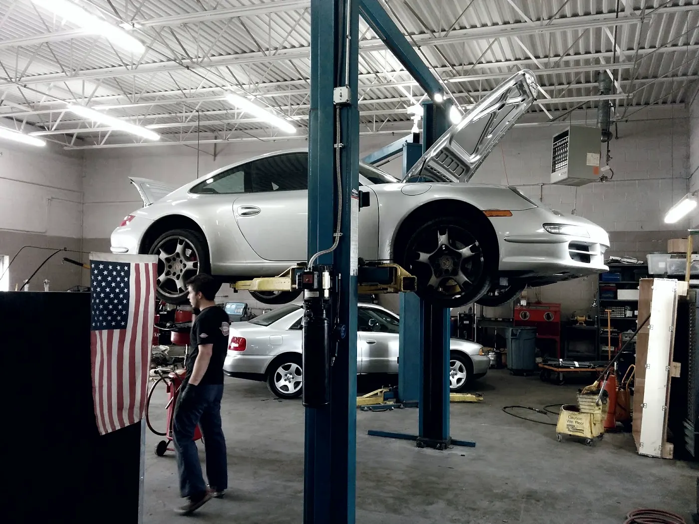 A silver Porsche 911 raised on a lift in the service bay, an Audi parked behind it.